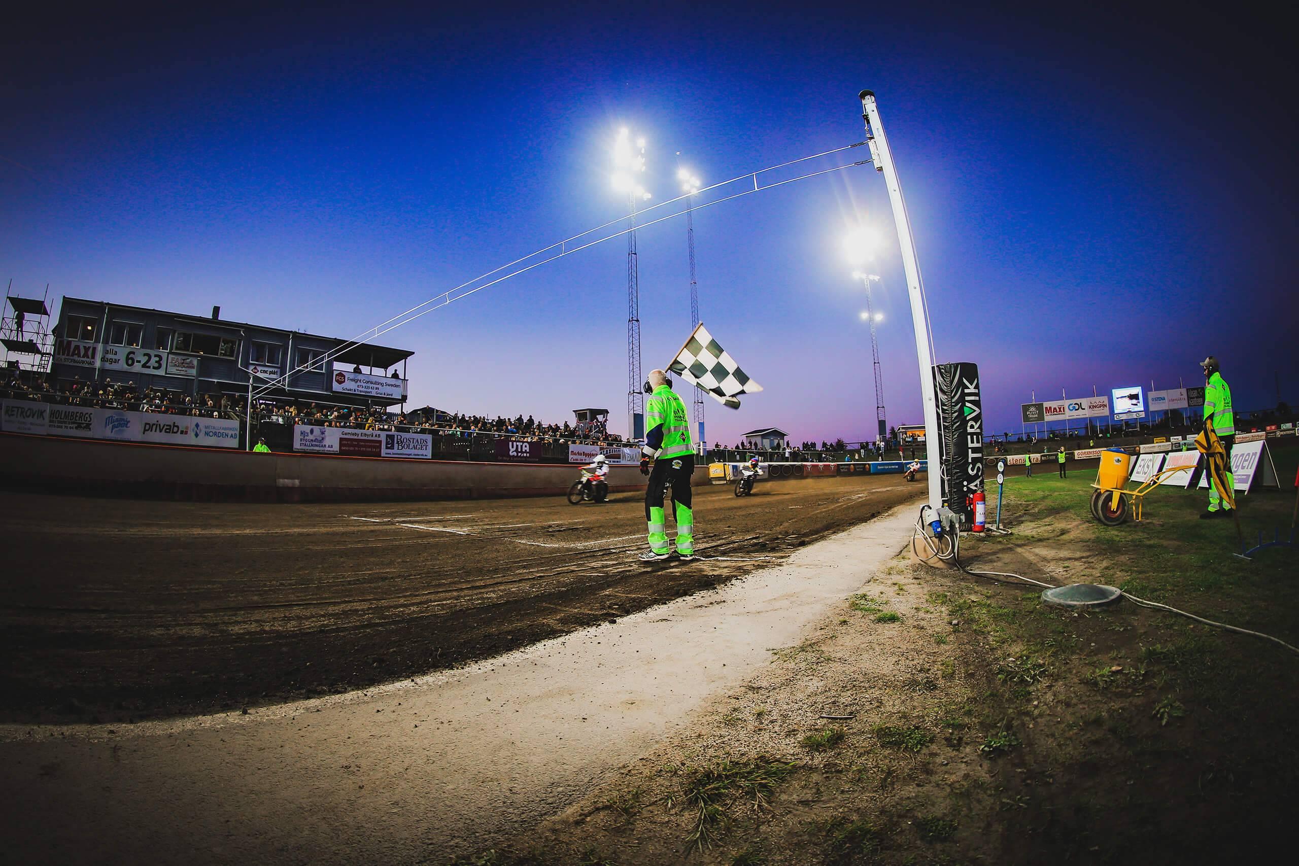 En funktionär i gröna varselkläder håller upp en målflagga när två speedwayförare kör i mål.