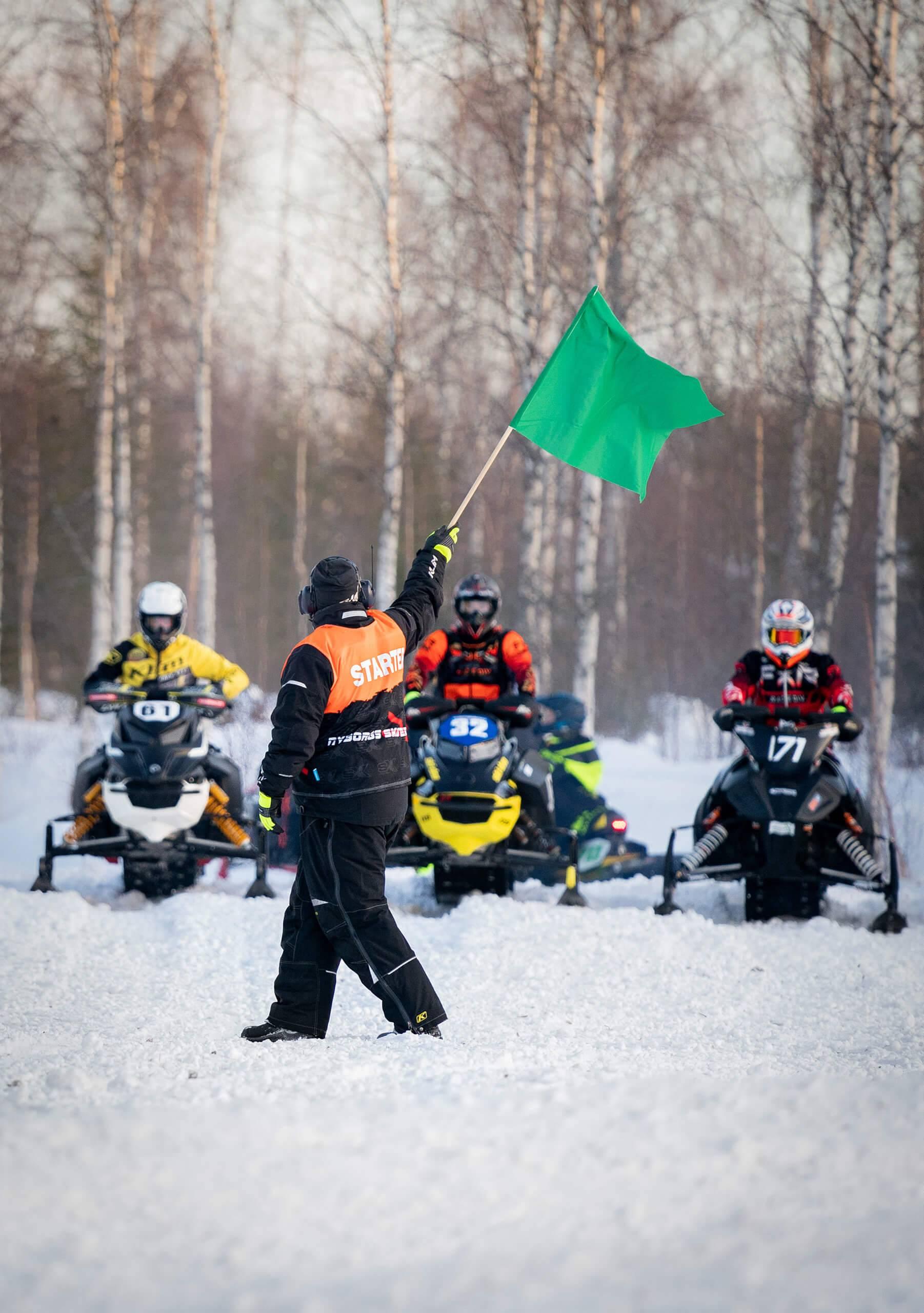 En Svemofunktionär håller upp en grön flagga framför skoterförare vid startlinjen.