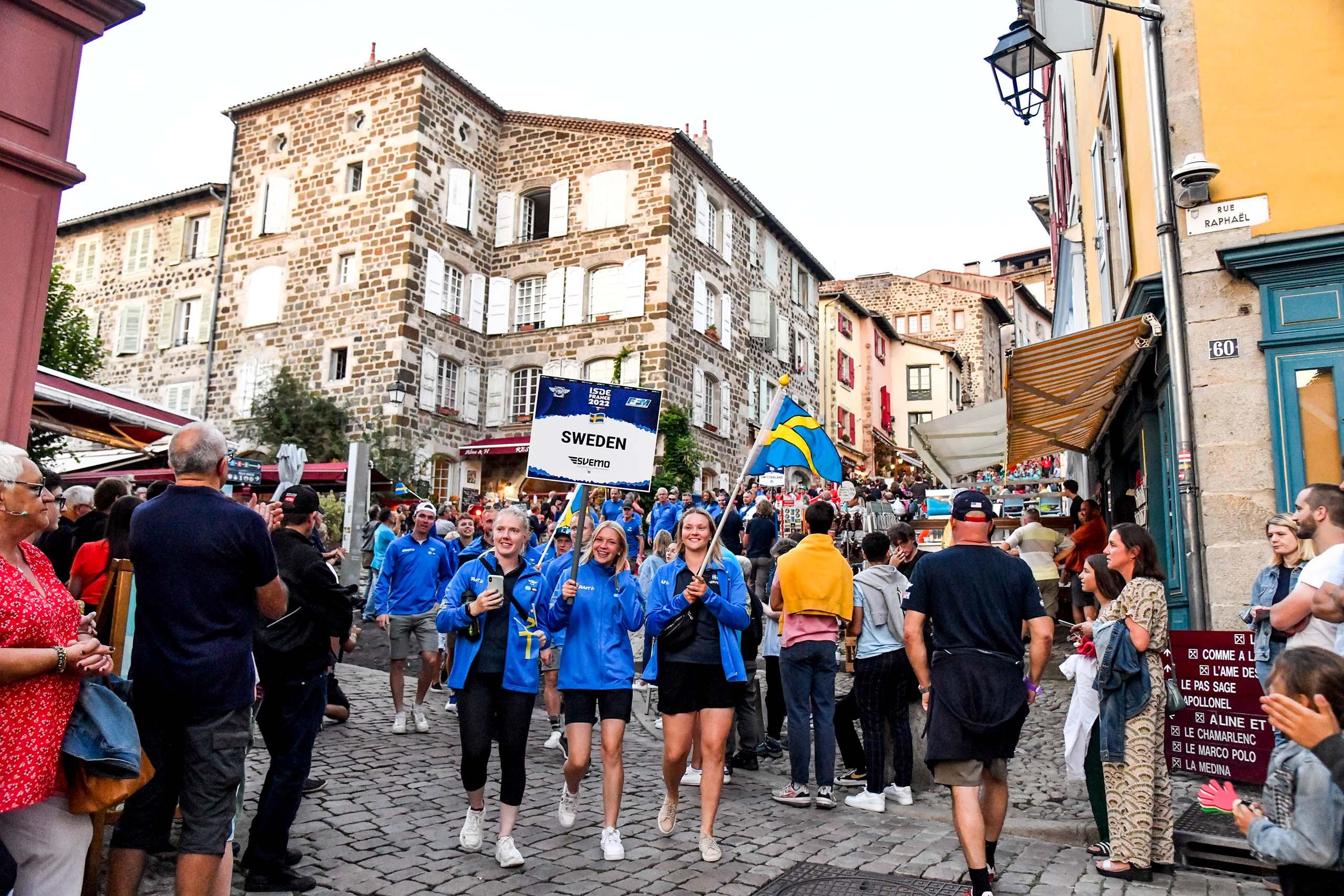 Många glada personer i blå-gula kläder på gatan håller upp en skylt som det står Sweden Svemo på, och viftar med den svenska flaggan