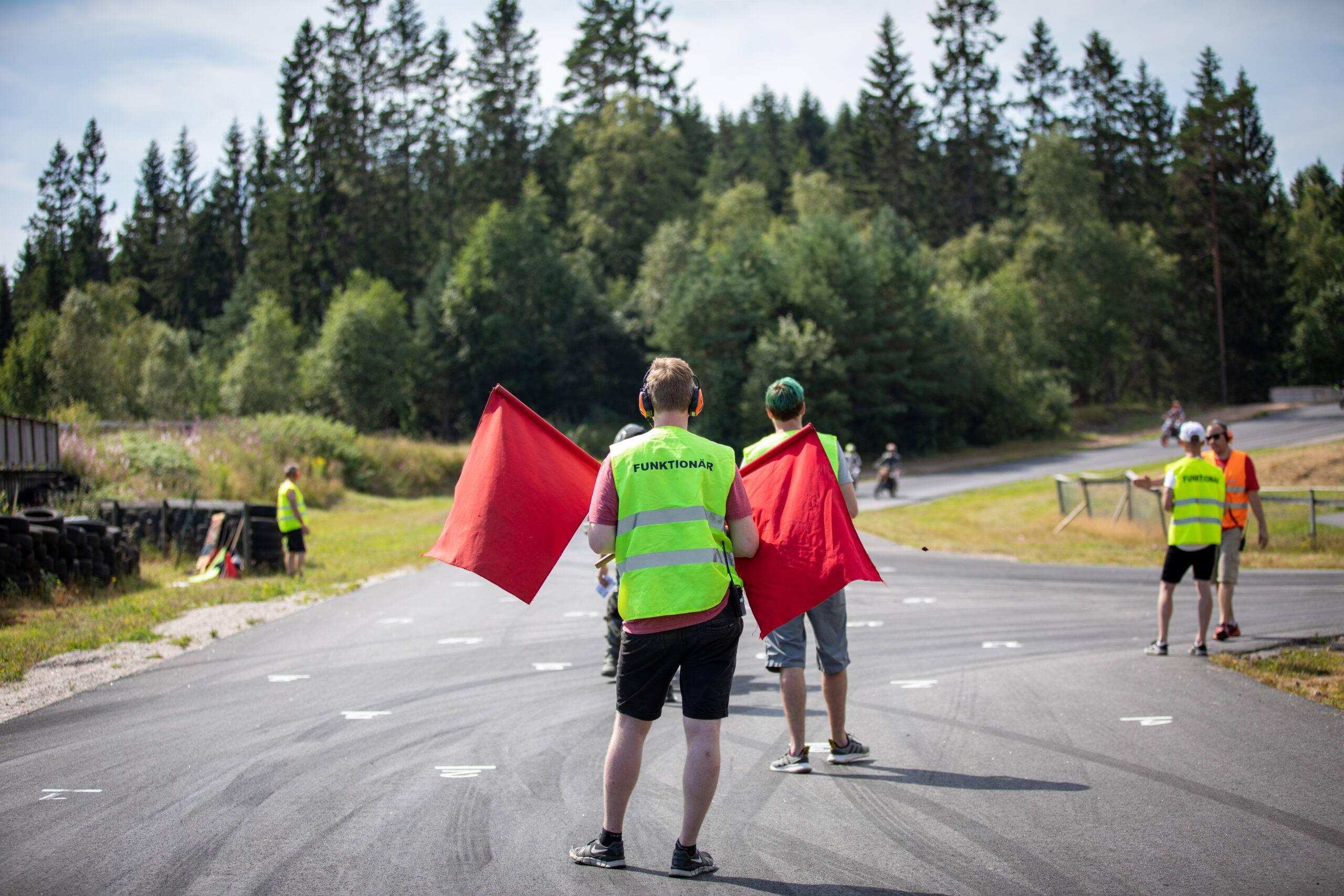 En funktionär står på en asfaltbana och håller två röda flaggor. Runt honom på banan står fler funktionärer.