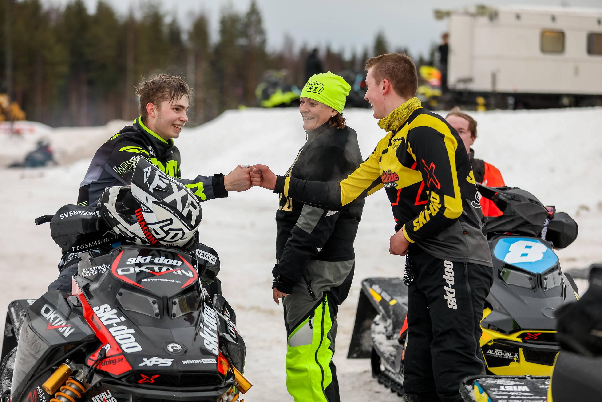Två glada och svettiga snöskoterförare som tagit av sig hjälmarna slår knytnävarna ihop med varandra efter en tävling. En lika glad mamma ser på.