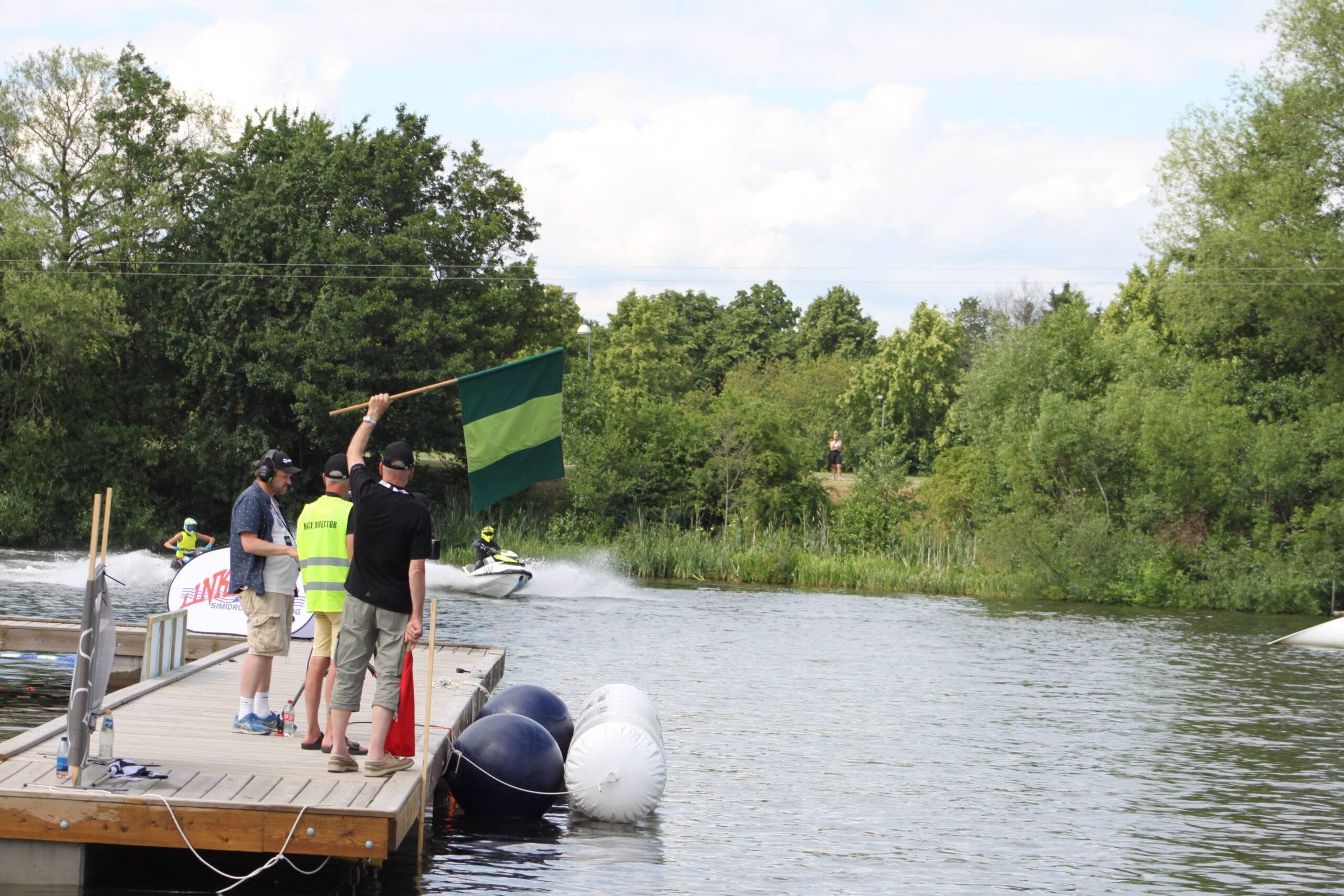 Två funktionärer står på en brygga och håller ut en grön flagga