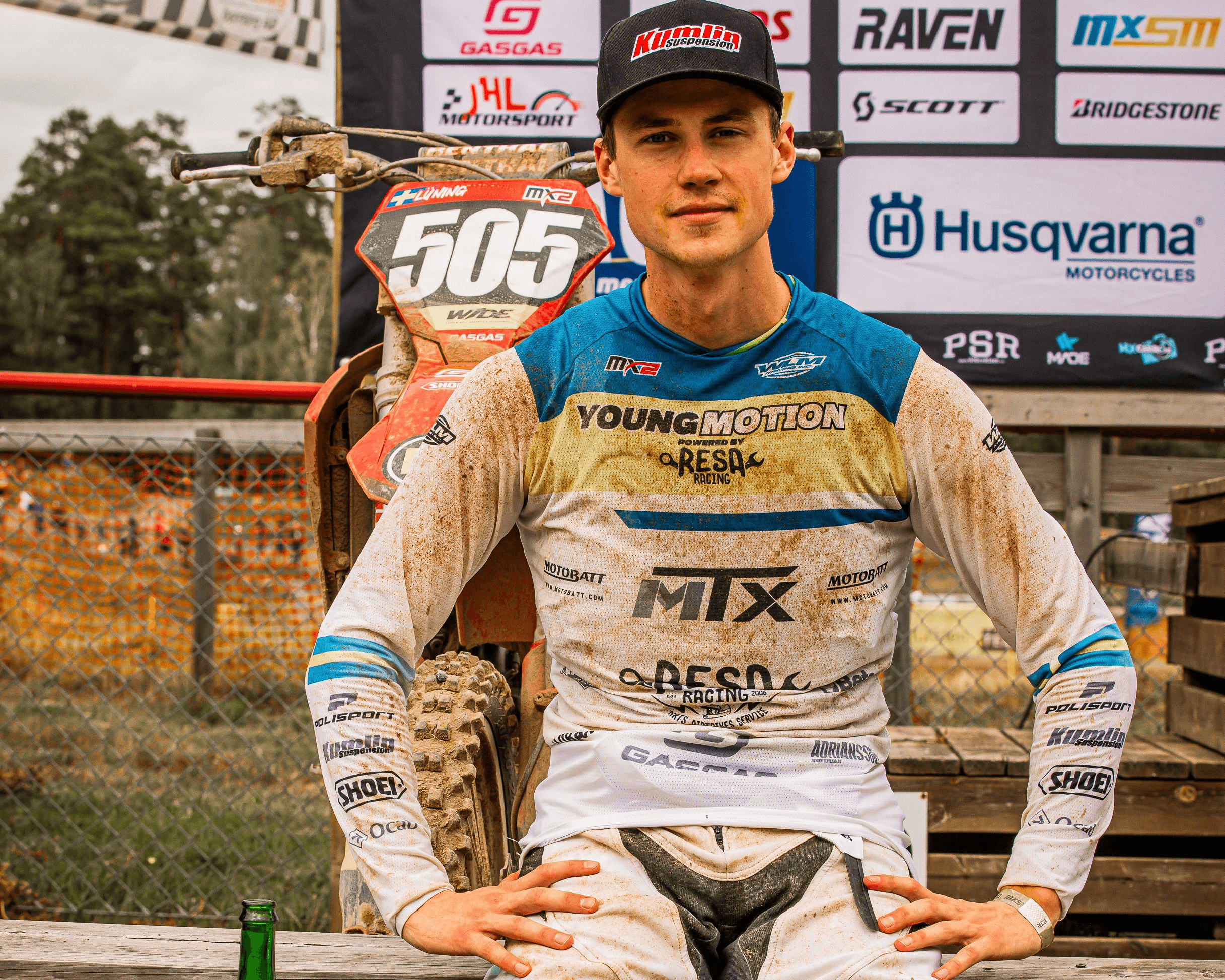 Arvid Lüning sitter på ett staket i leriga motocrosskläder med sin hoj bakom sig och en flaska pommac framför sig. I bakgrunden sysn en crossbana