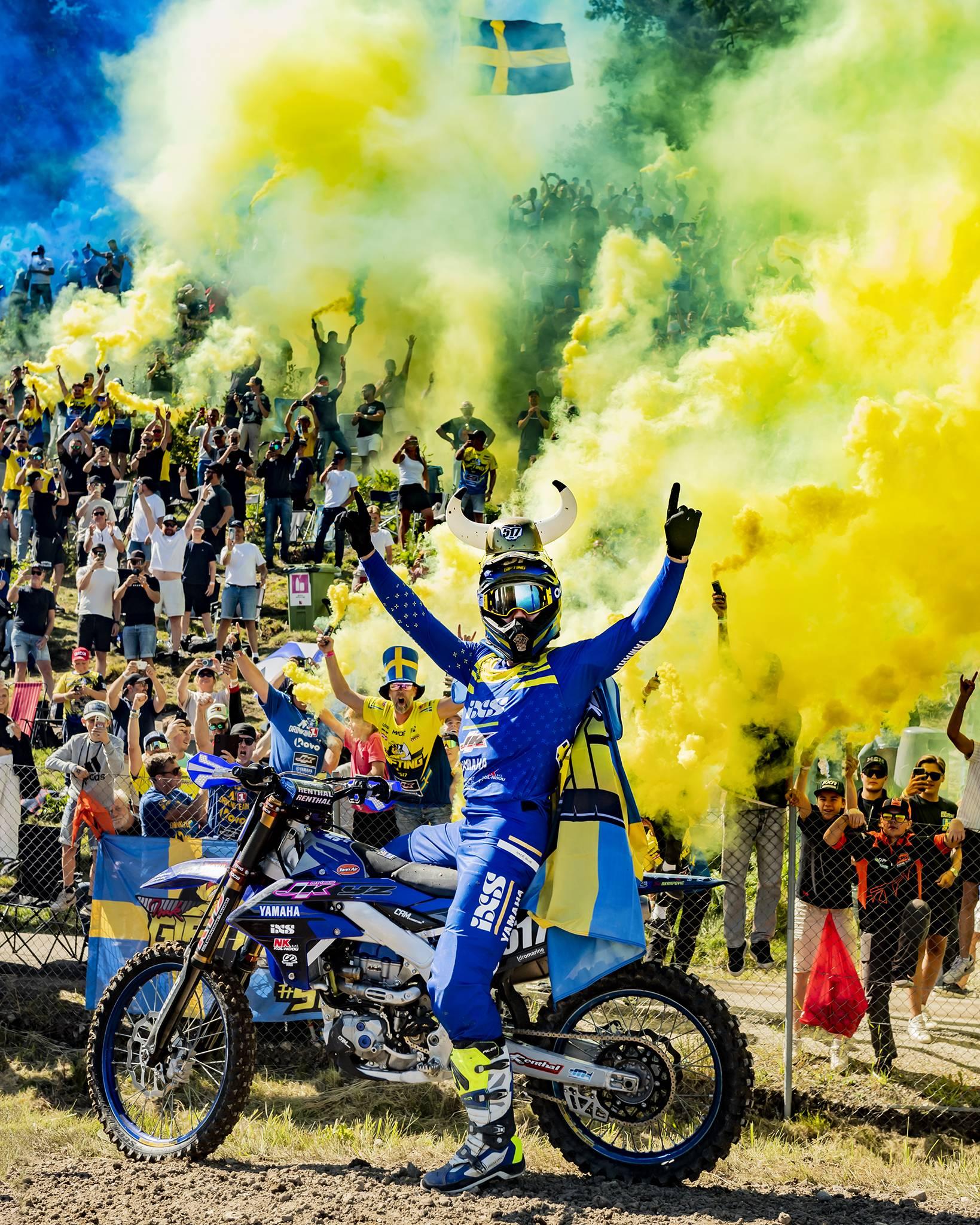 En bild på SM segraren i motocross 2024 Isak Gifting. Han sitter på sin motocross och håller upp båda armarna i luften i en segergest, på huvudet har han en hatt med vikinghorn. I bakgrunden ett punlikhav med supportrar som hejar och ropar ut sin segerglädje. Bengaler färgar hela publikhavet och himmelen blå och gul.