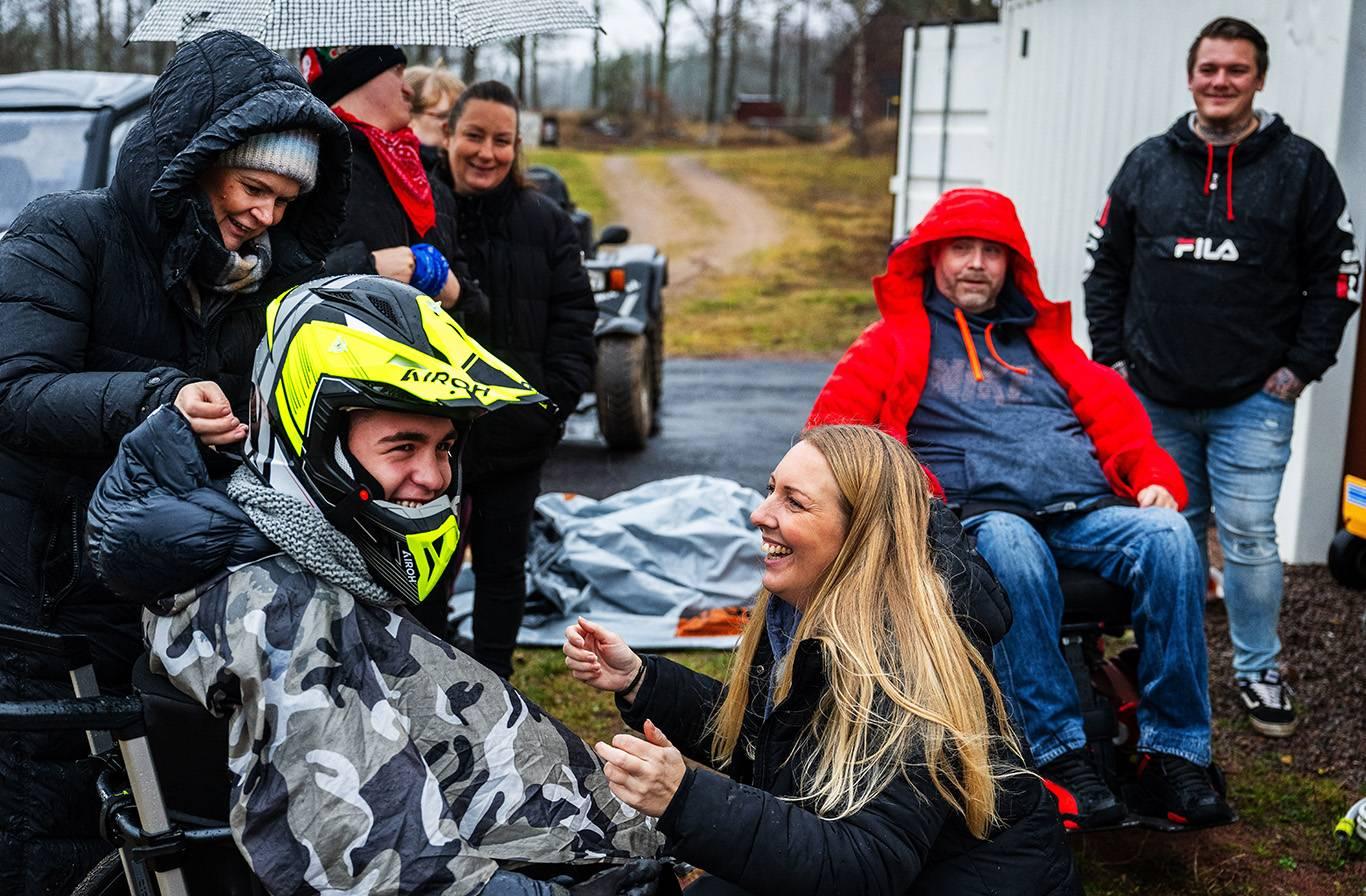 Ett gäng med motorsportsutövare och föräldrar har samlats på en motorbana för att utöva motorsport anpassade efter funktionshindrade. Två personer sitter i rullstol och en av dem gör sig redo att köra med hjälm och skyddsutrustning på sig. Med sig har de människor som är glada och hjälper till.