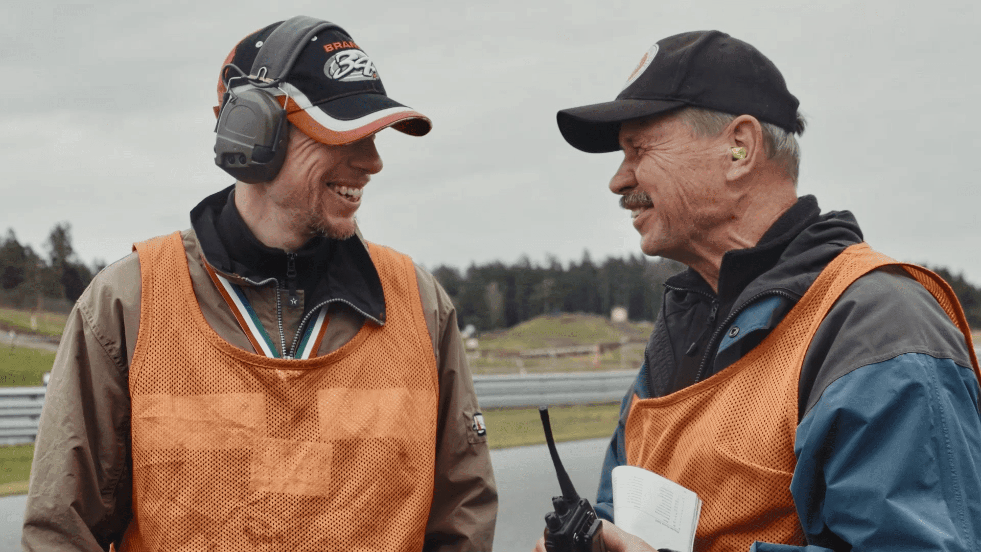 Två äldre herrar tittar på varandra och skrattar tillsammans klädda i orangea funktionärsvästar, keps och hörselkåpor.