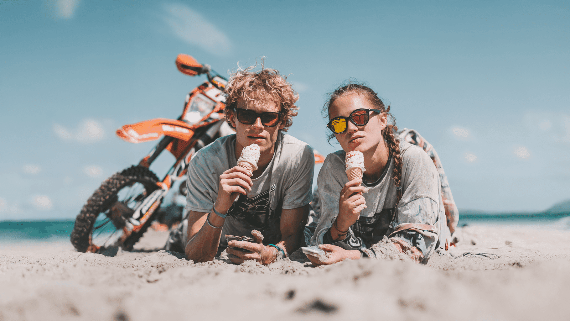 Två unga enduroåkare ligger på en solig strand och äter varsin glass. Endurocyklarna står i bakgrunden